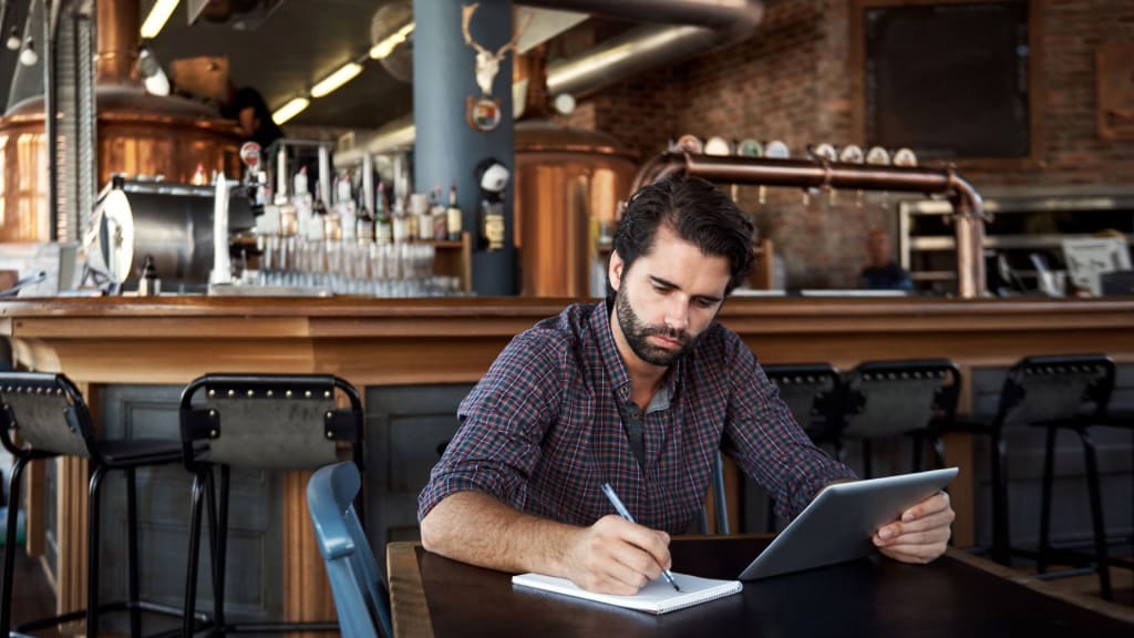 A man sits in a restaurant or bar, writing in a notebook and looking at a tablet, possibly researching types of liquor licenses in California. The background includes empty bar stools, taps, and brewing equipment.